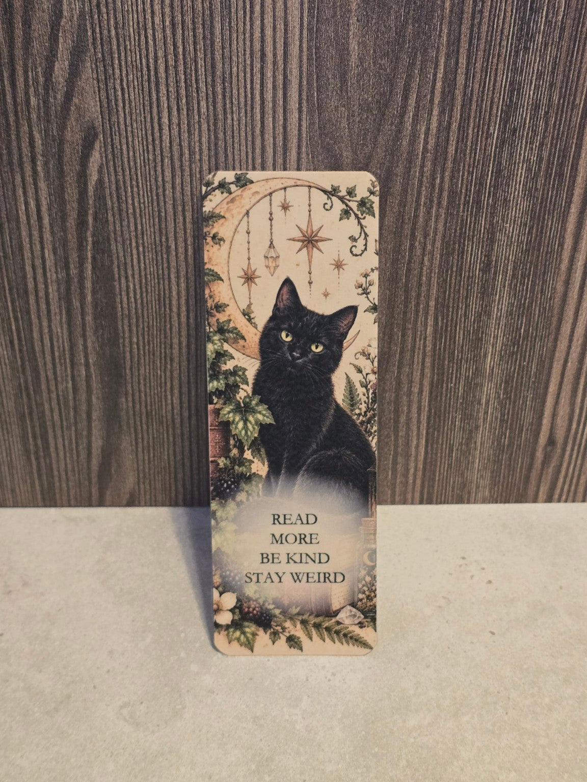 Read More Be Kind Stay Weird laminated bookmark with a black cat, golden crescent moon and woodland plants.
