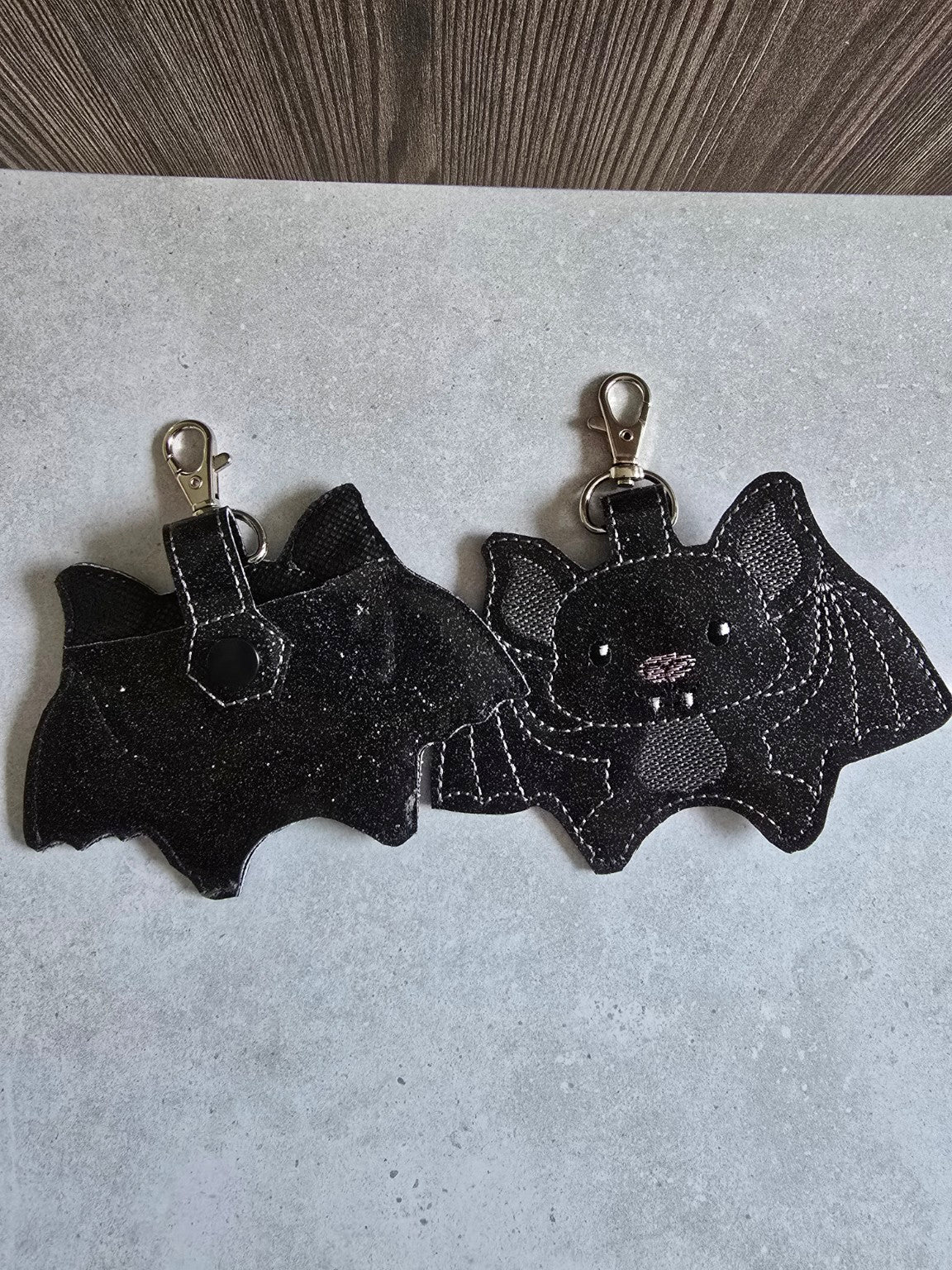 Back and front of embroidered bat sanitizer holders, showing the snap-closure tab and metal keychain clips.