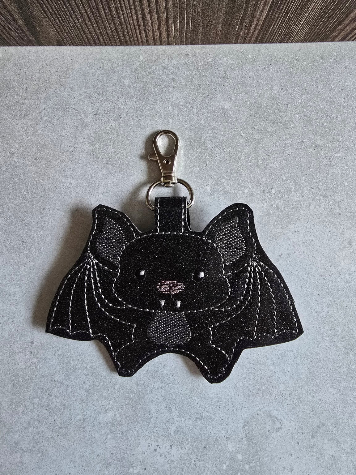 Front view of a black embroidered bat hand sanitizer holder with silver stitched wings, a pink nose, tiny fangs, and a silver swivel clasp.