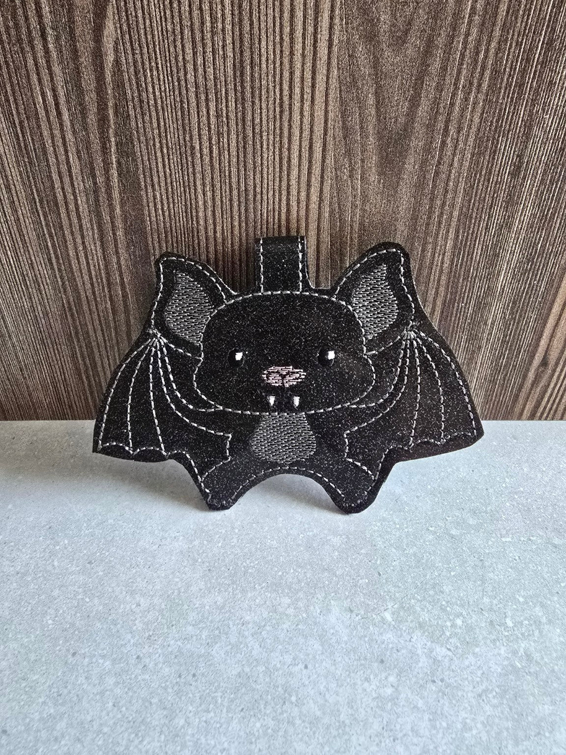 Black sparkly bat hand sanitizer holder standing upright, showing its embroidered wings, face, and hanging tab.