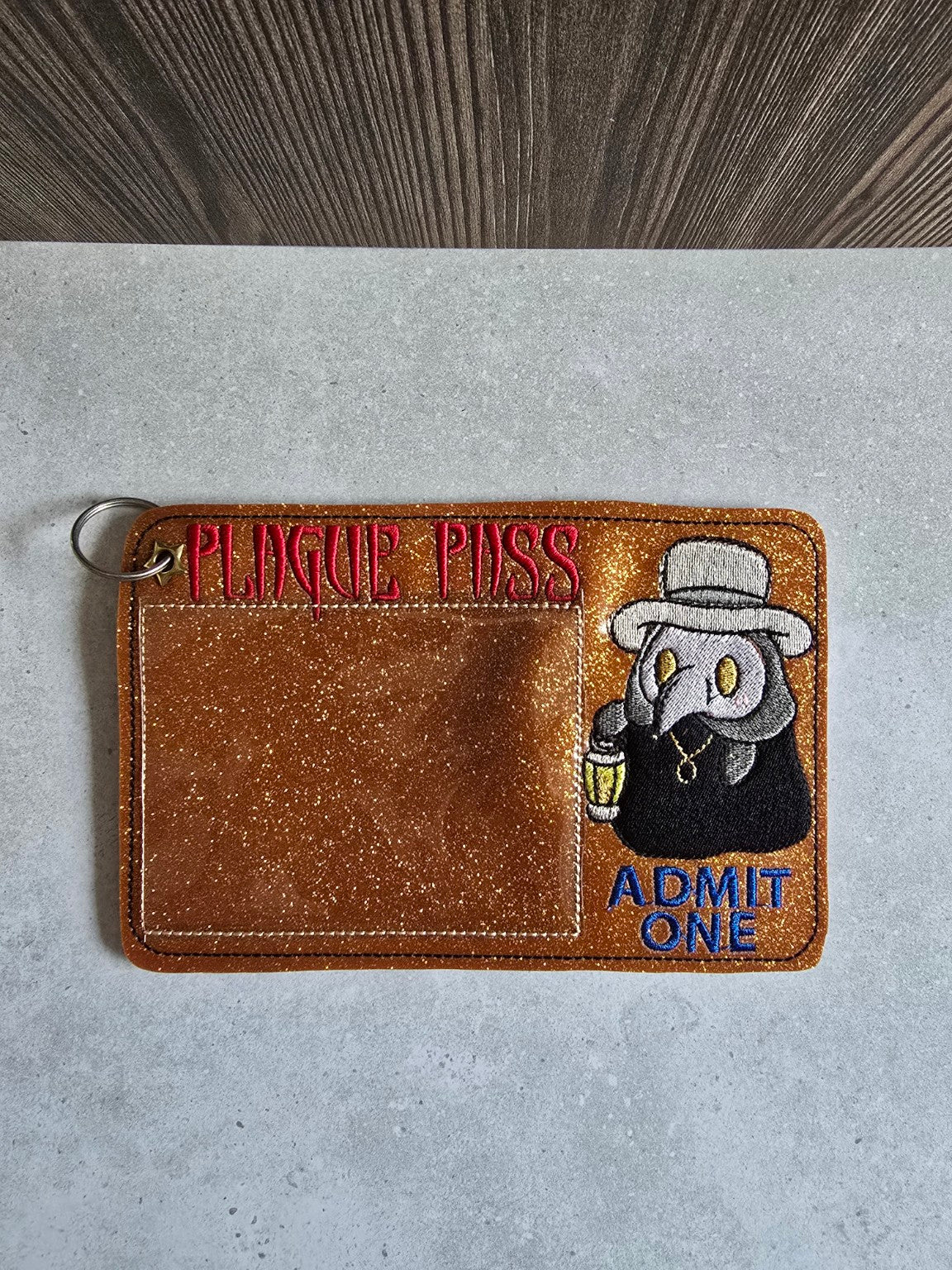 Front of the copper-orange glitter embroidered Plague Pass holder with clear card pocket, plague doctor holding lantern and Admit One lettering.
