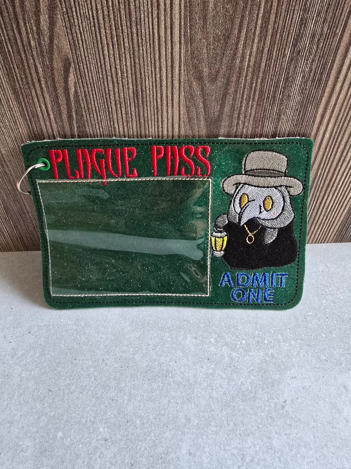 Front of forest-green glitter Plague Pass embroidered holder, showing clear card pocket, plague doctor embroidery and metal ring.