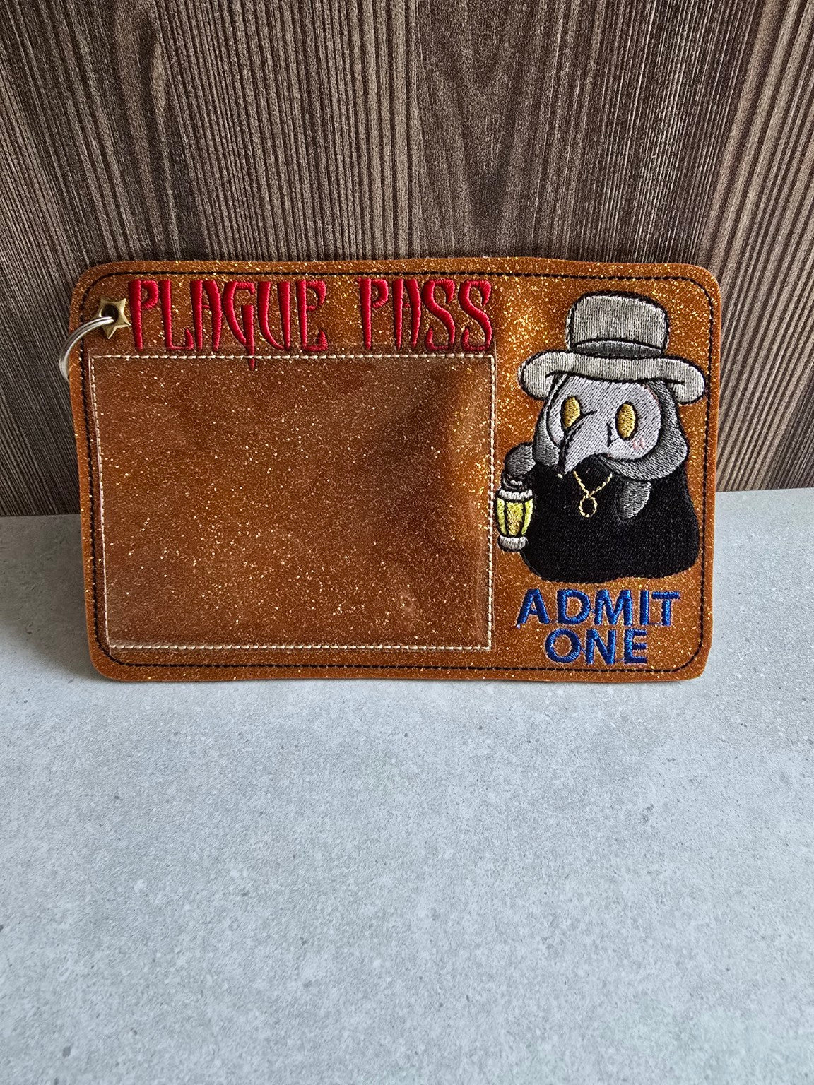 Forest-green glitter plague doctor pass holder standing upright with clear card pocket, red Plague Pass and blue Admit One lettering.