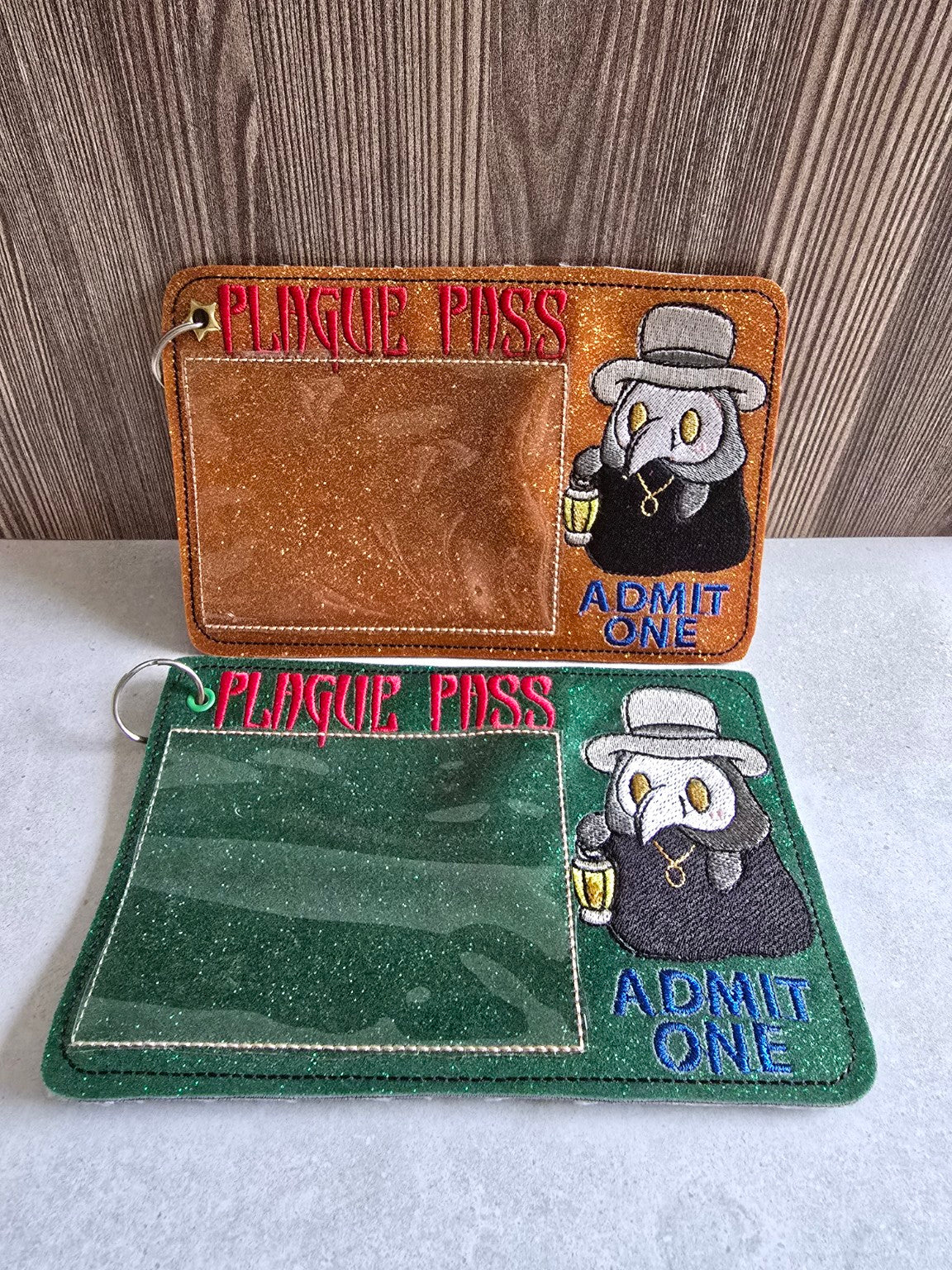 Two final embroidered plague doctor pass holders shown together: copper-orange glitter and forest-green glitter versions.
