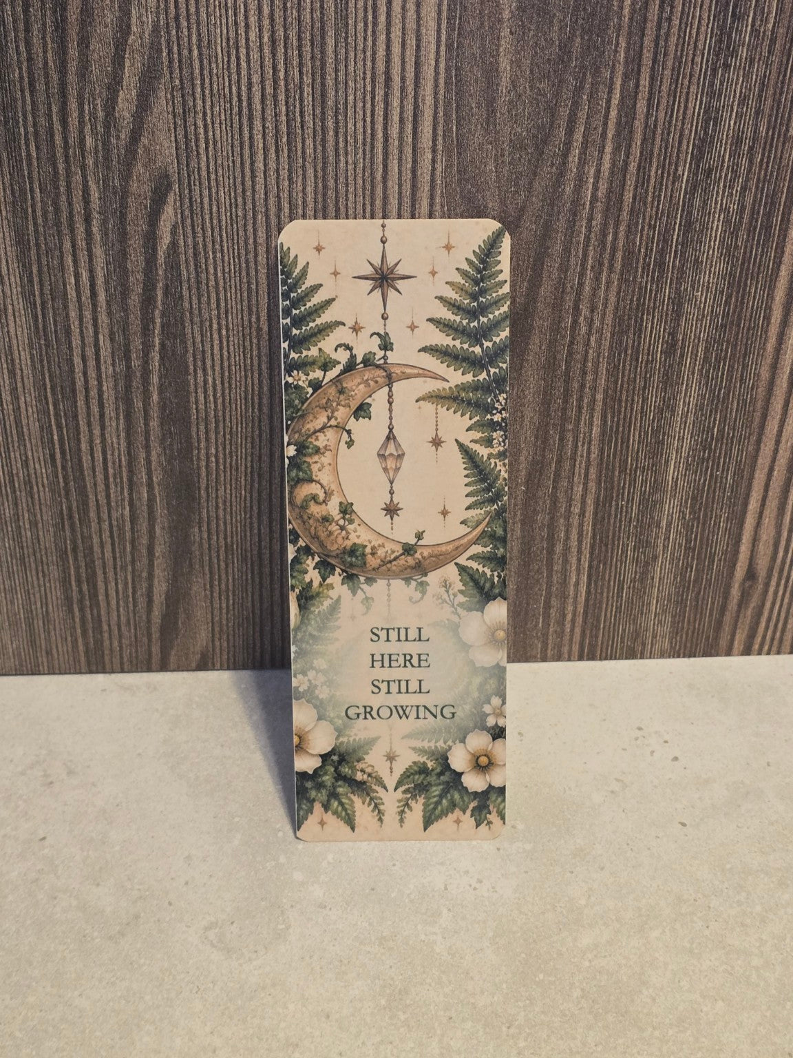 Still Here Still Growing laminated bookmark with a crescent moon, hanging crystal, ferns and white flowers.