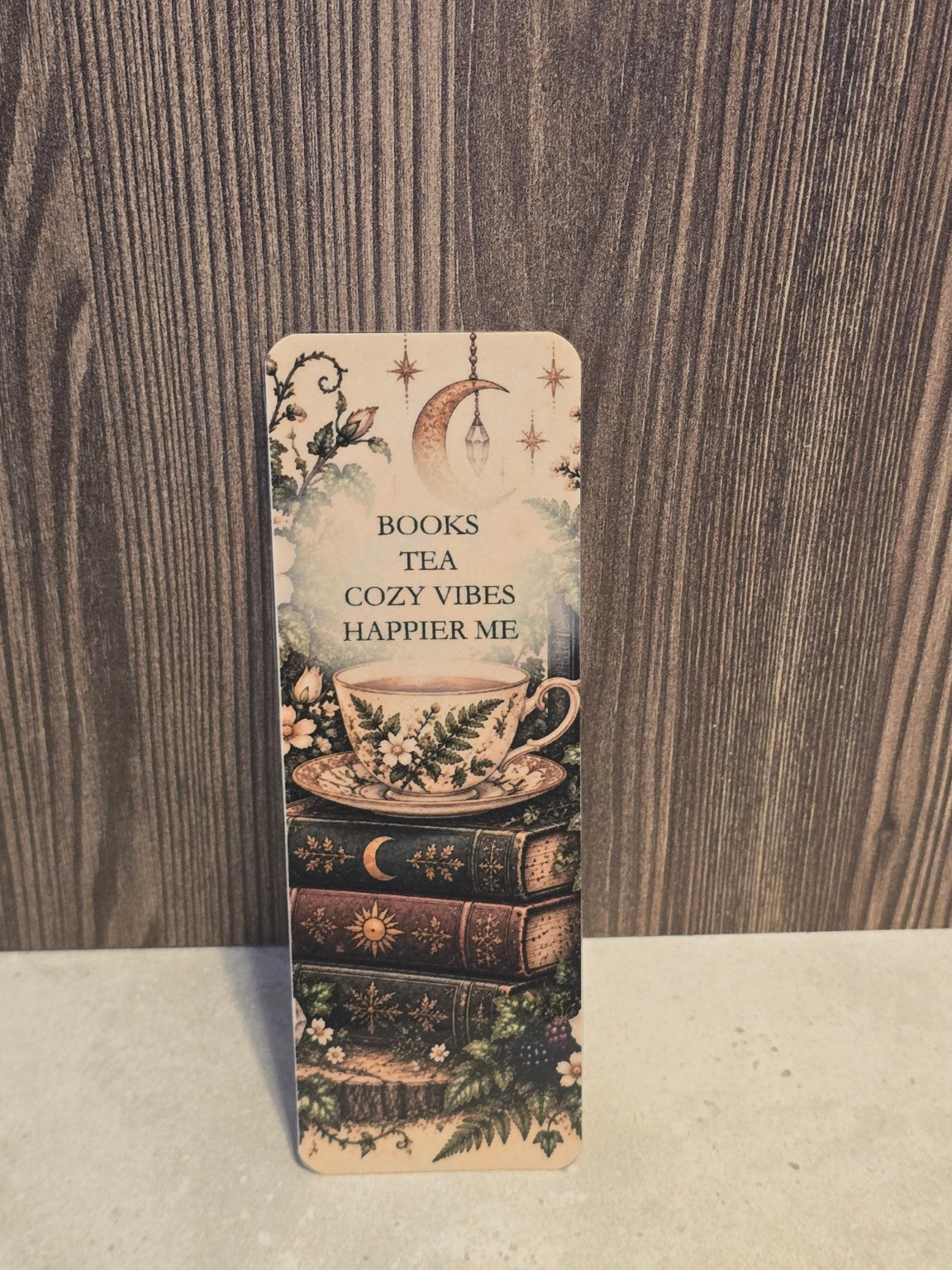 Books Tea Cozy Vibes Happier Me laminated bookmark showing a floral teacup on antique books with moon and greenery.