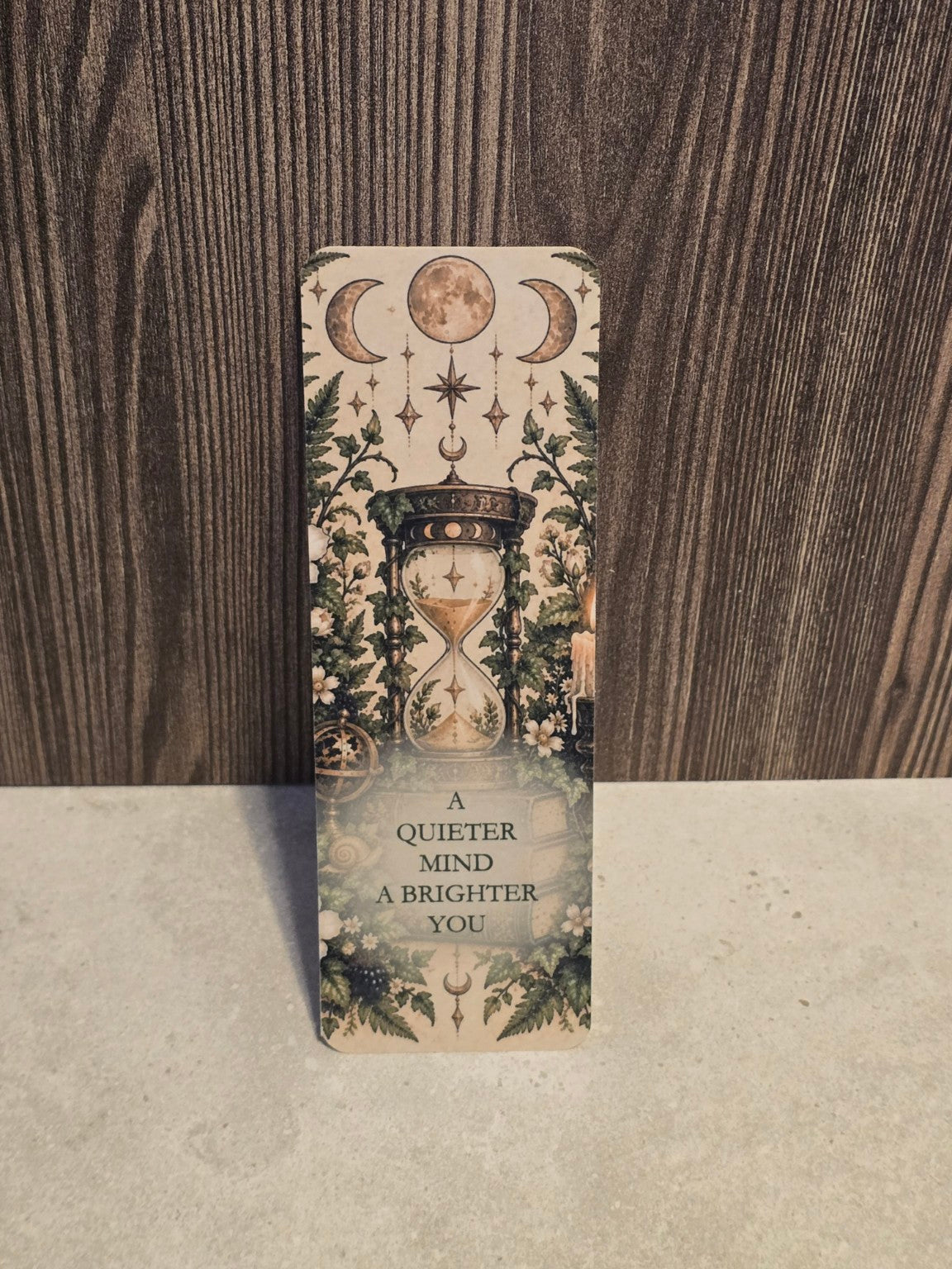 A Quieter Mind A Brighter You laminated bookmark with an ornate hourglass, moon phases and woodland foliage.