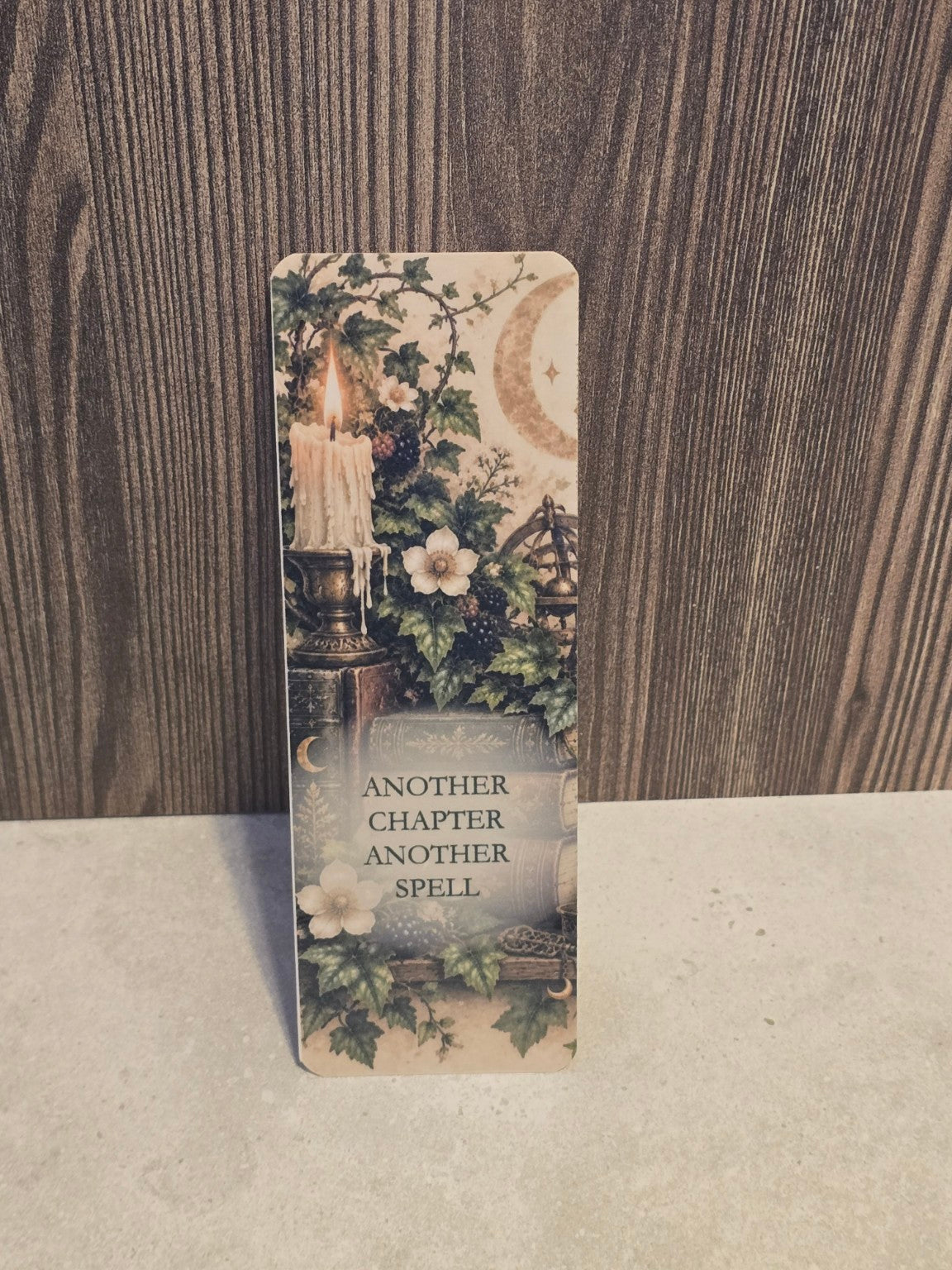 Another Chapter Another Spell laminated bookmark featuring a lit candle, antique books, crescent moon and flowers.